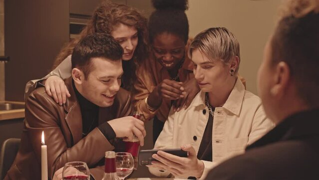 Medium slowmo of feminine gay man with dyed blonde hair showing photos on smartphone to his LGBTQ friends while having small cozy dinner party in kitchen with dimmed lights