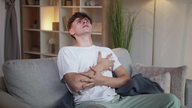 Heart pain. Agony man. Cardiac stroke. Young guy in casual look grasping chest feeling unbearable injury sitting sofa light home interior.