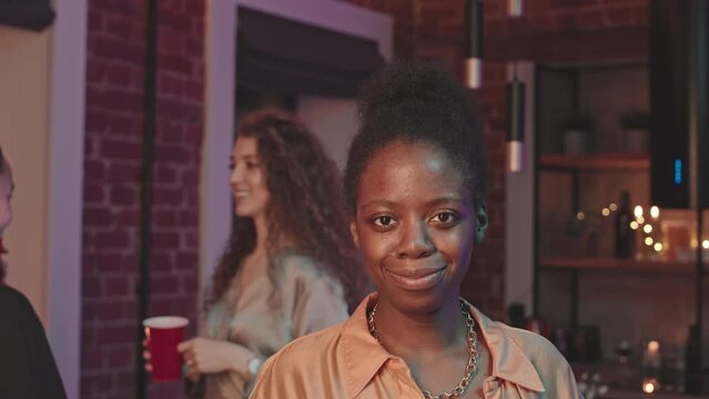 Waist up slowmo portrait of young beautiful Black woman smiling at camera during home party while her female friends with plastic red cups in hands chatting in background