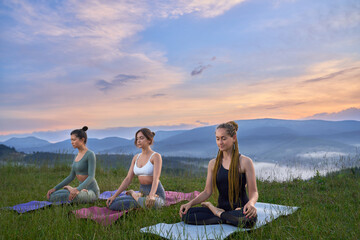 Fototapeta na wymiar Three beautiful women with closed eyes meditating on yoga mat in lotus pose, deeply breathing and smiling. Pretty female among grass relaxing after yoga practice at sunrise time. Concept of balance.