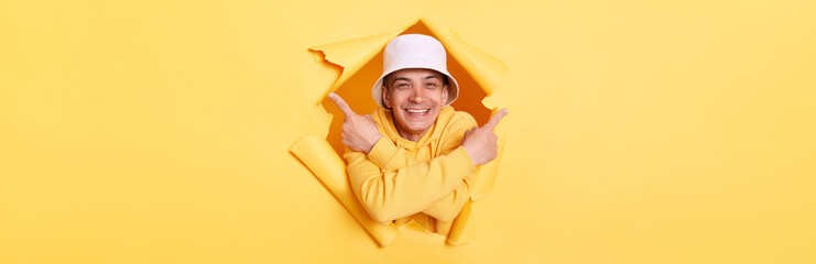 Portrait of smiling satisfied man wearing casual hoodie and panama looking through torn hole in yellow paper, pointing with both index fingers, showing area for your advertisement.