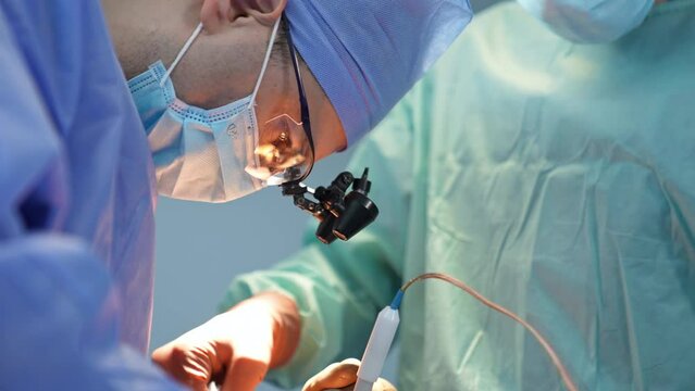 Chief surgeon in mask, cap and device glasses bent his head low over the patient. Close up portrait of a specialist focused on surgery.
