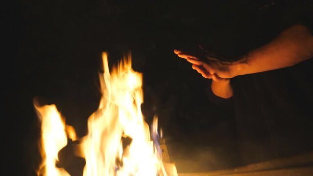 Low view of young tourist rubs his hands in front of an open bonfire from mangal at night. Man warms his arms by a fire from metal brazier against the black background. Concept of warmth and tranquil