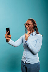 Beautiful confident young lady greeting friend over online phone video call while standing on blue background. Happy smiling african american woman with smartphone waving at video conference call