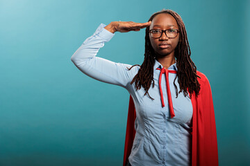 Serious and confident superhero woman standing on blue background while saluting patriotic. Brave...