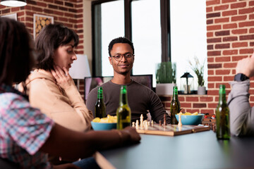 Smiling man sitting at table in living room with friends while watching a game of chess. Intelligent multiethnic people playing strategy boardgames while enjoying snacks and beverages.