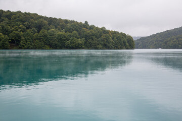Plitvice Lakes National Park, Croatia, Europe: Lake with beautiful turquoise and calm water reflecting the dark green forest on a cloudy and foggy morning