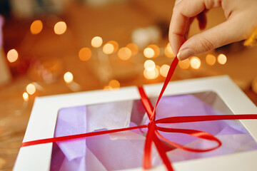 Close-up of girl's hand pulling ribbon from surprise box. Bokeh lights in background. Unpacking gift on wooden table. Festive concept of Christmas or Birthday.