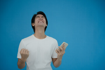 Cool asian man excited and surprised by smartphone on blue background. A man saw a sale on an online shopping website or See the results of the football team cheering competition.