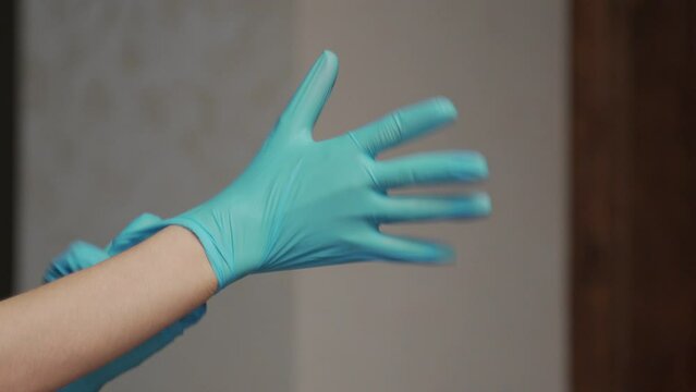 Young woman puts on blue rubber gloves on hands on blurred background in room. Housewife gets prepared for cleaning dust in apartment close view