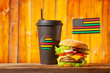 Classic American burger with alternative African American juneteenth flag on the top and black paper cup with straw over wooden background. Close-up with selective focus.