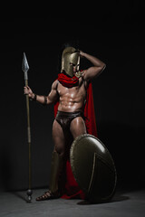 A young athletic sexy man dressed as a Roman warrior in a red cloak stands with a spear in his hand...