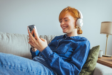 A stylish young woman is sitting on a sofa and listening to music with a wireless bluetooth...