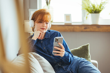 Shot of a young woman using a smartphone and headphones on the sofa at home. Stylish young woman listening to music at home. A woman is sitting on a sofa and listening to music