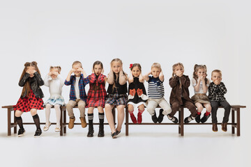 Little girls and boys, funny children sitting on banch isolated on grey studio background. Beauty, kids fashion, education, happy childhood concept.
