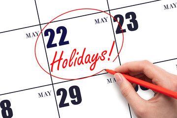 Hand drawing a red circle and writing the text Holidays on the calendar date 22 May. Important date.