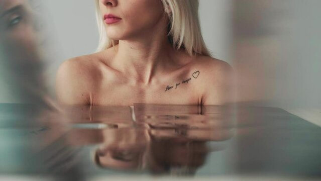 Tattoo woman. Body art. Feminine beauty. Attractive elegant young female with bare shoulders romantic writing on collarbone at blurred mirror glass reflection.
