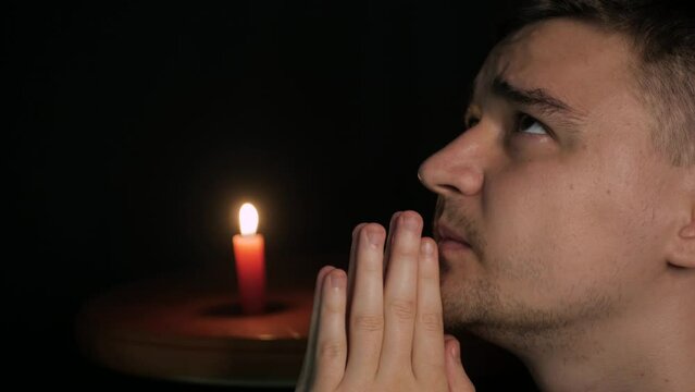 Close-up of young caucasian male praying to God with yellow candle in background. Sinner folding hands together, looking up and saying a quiet prayer to Jesus, begging for forgiveness for his soul. 
