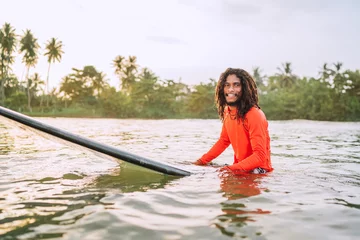Wandaufkleber Black long-haired teen man floating on long surfboard, waiting for a wave ready for surfing with palm grove litted sunset rays. Extreme water sports or traveling to exotic countries concept. Sri Lanka © Soloviova Liudmyla