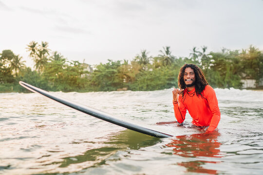 Black long-haired teen man showing shaka sign floating on long surfboard, waiting wave ready surfing with palms litted sunset rays. Extreme water sports or traveling to exotic countries concept