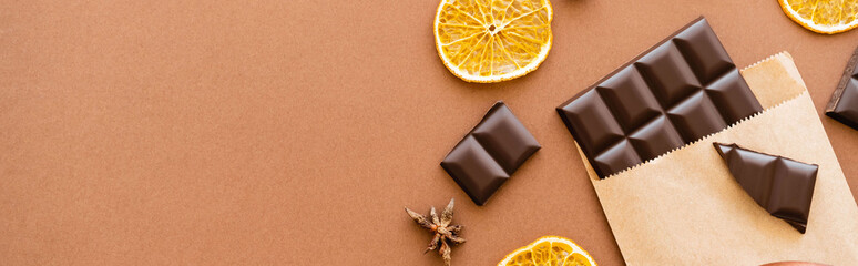Top view of anise, dry orange slice and chocolate in craft package on brown background, banner.