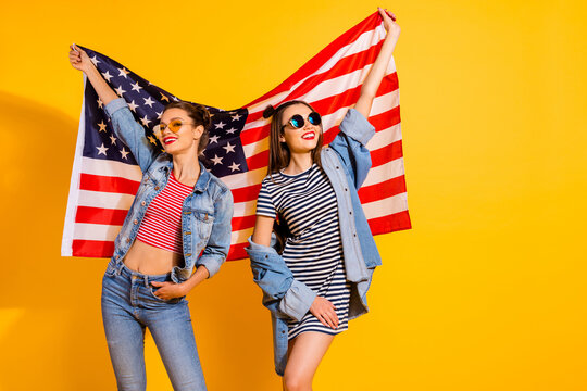 Portrait of two attractive positive girls hold american national flag isolated on yellow color background