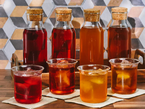 Four bottles and four glasses of cold brew fruit tea, rooibos, green and black, in a wooden tray on a wooden table
