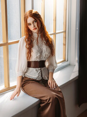 Red-haired woman in vintage dress sits on large classic window waiting love. Clothing costume...