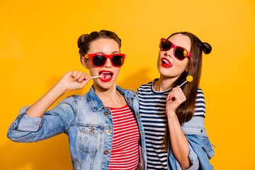 Photo of two funky positive girls lick lollipop candy enjoy free time isolated on yellow color background - obrazy, fototapety, plakaty