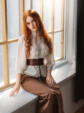 Red-haired woman in vintage dress sits on large classic window waiting love. Clothing costume countess old art style white blouse, brown long skirt. Curly red hair. Redhead girl princess 1800s stylish