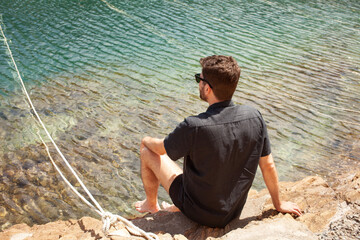 Man sitting from behind next to the water and enjoying sun and beautiful transparent Mediterranean blue sea and coast line. Wearing casual clothes linen shirt and sunglasses. Spanish summer scene