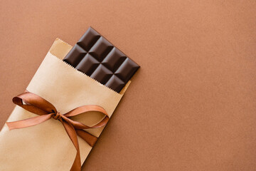 Top view of dark chocolate bar in craft package with ribbon on brown background.