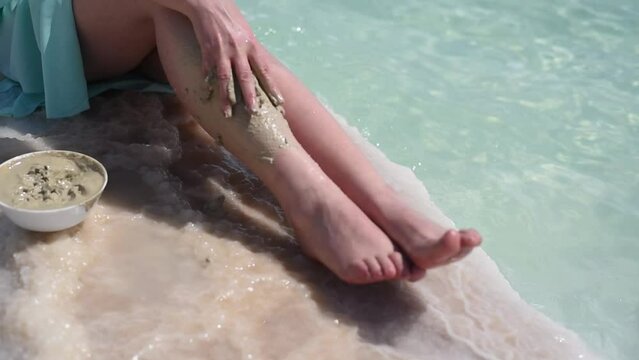 Woman's legs. Person sits on white salty beach near water of Dead sea and by hand puts gray mud on leg. Healthy vacation on Dead Sea with mineral mud. Travel and holiday in summer
