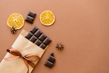 Top view of anise and orange slices near chocolate in craft package with ribbon on brown background.