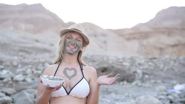Smile girl in hat and white bikini holds bowl with gray mud on rocky shore background. Happy vacation on Dead Sea with therapeutic mud on face and body. Travel to hydrogen sulfur hot water in Israel