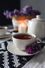 A cup of tea with a branch of lilac on a saucer on the table. Candle, teapot, wicker basket with lilacs. Aesthetics of tea drinking