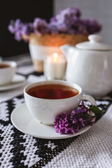 Obraz na płótnie Canvas A cup of tea with a branch of lilac on a saucer on the table. Candle, teapot, wicker basket with lilacs. Aesthetics of tea drinking
