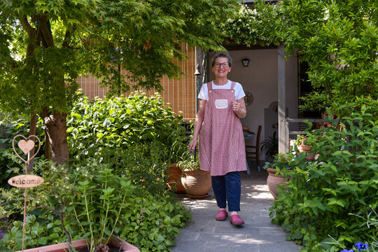 60 Year Old Woman In Her Home Garden Looking At The Plants, Wide Shot Looking At The Camera