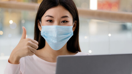 Asian korean successful satisfied business woman worker freelancer student girl designer specialist in face medical mask with laptop looking at camera showing thumb up recommends wifi computer app