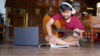 A cute Asian boy wearing headphones video calls learning  .music and writing songs online on his...