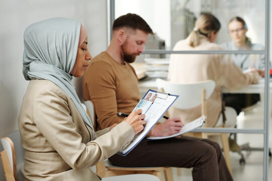 Young adult Caucasian man and Muslim woman in hijab sitting on chairs in visa service agency filling in application forms while waiting for appointment