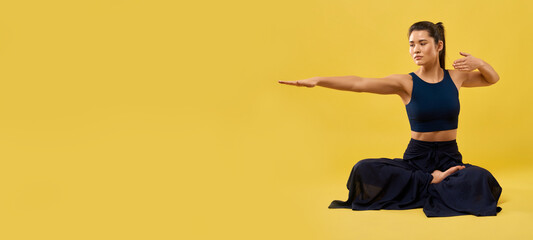 Pretty girl holding right arm outstretched, while sitting in meditation pose indoor. Front view of lady practicing yoga, keeping one hand bent, isolated on orange studio background. Concept of yoga.