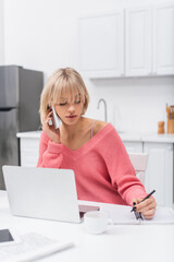 young blonde woman talking on smartphone while writing near laptop.