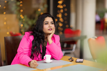Portrait of a young and stylish Indian woman sitting in a cafe and enjoying a drink while waiting for friends, colleagues or customers