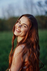 Naklejka premium Portrait of a woman with a beautiful smile in summer green nature against the grass, looking out at the setting sun. The concept of health and beauty