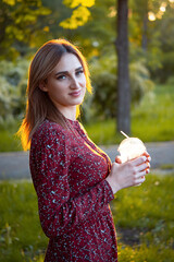 woman walking on a park.She drinks coffee from "to go" cup. Stunning sunset on a background.A young blonde girl walks in the park. She is wearing a red midi dress and white sneakers. Lai stayed.