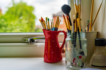 Kolekcja pędzli do malowania w czerwonym wazonie stoją na oknie. © Aneta