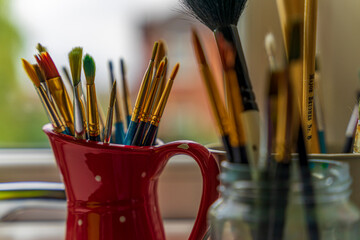 Kolekcja pędzli do malowania w czerwonym wazonie stoją na oknie. © Aneta