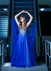 Beautiful blonde woman in a fluttering evening blue dress with a moving staircase on the background