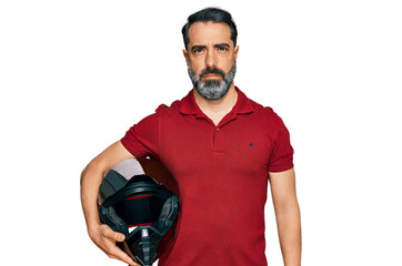 Middle aged man with beard holding motorcycle helmet thinking attitude and sober expression looking...
